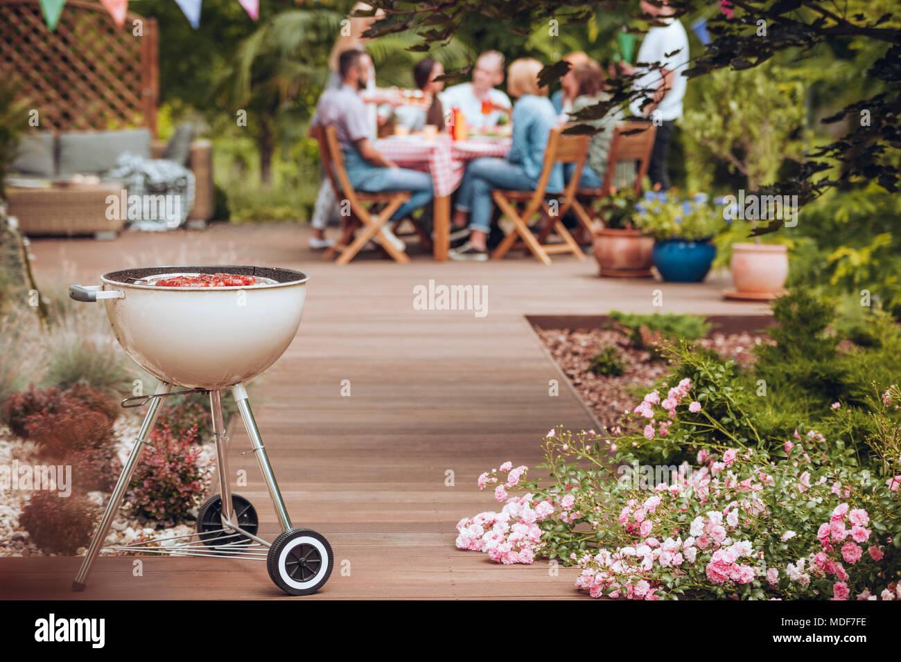 Grill with sausages in the garden with flowers and group of friends celebrating in the background Stock Photo