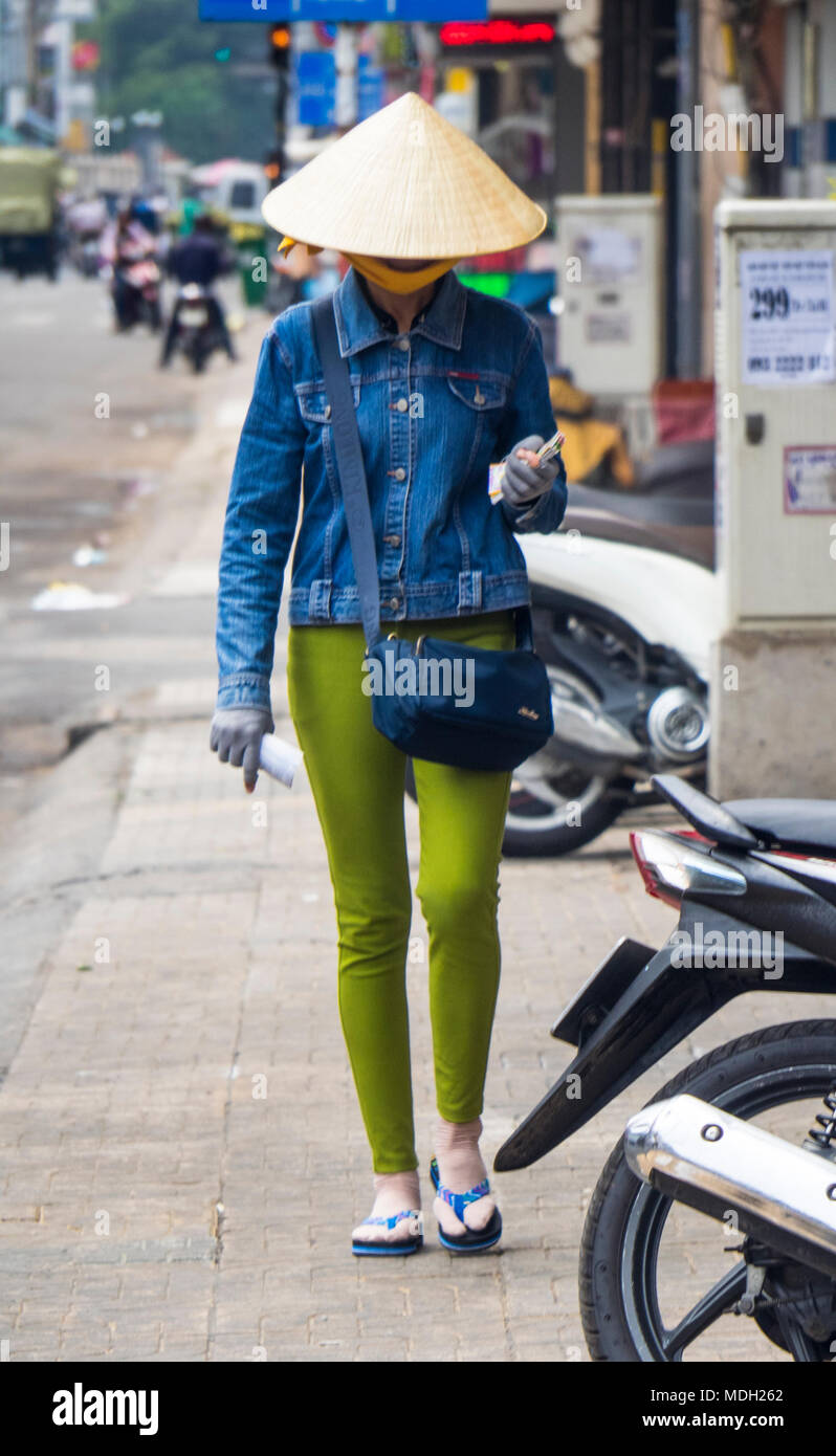 A Vietnamese woman wearing a conical hat, denim jacket and green trousers walking on the pavement on a street in Ho Chi Minh City, Vietnam. Stock Photo