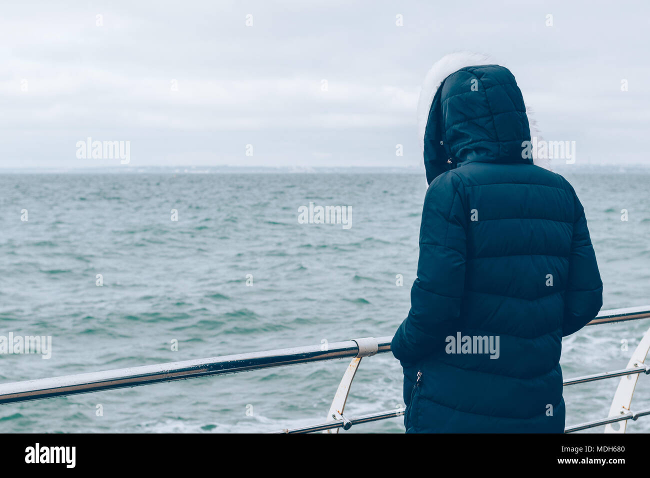 Rear view woman in blue down coat and hood looking at ocean on grey ...