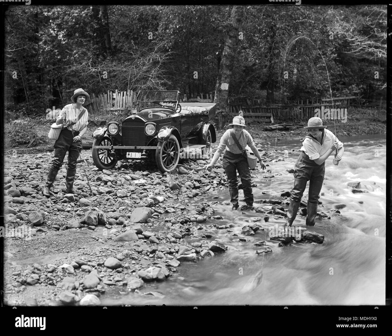 View of three pretty girls fishing in mountain stream with their 1919 'Studebaker' convertible automobile in the background. Studebaker sign displayed in windshield. Location in the mountains of California. Stock Photo