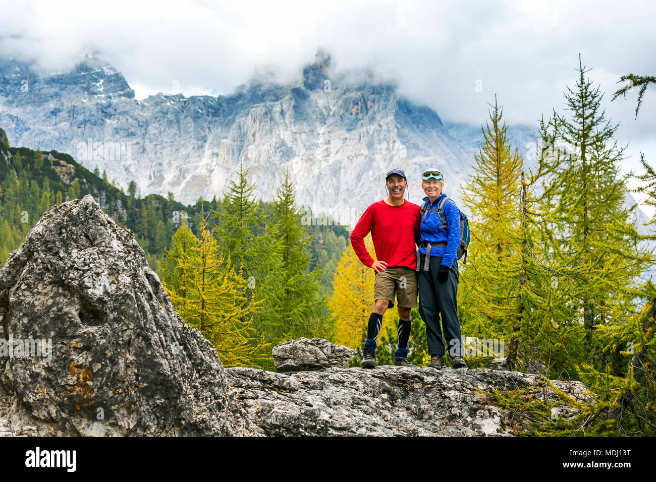 Male and female hikers standing on rock with autumn coloured trees and cloud-covered mountain range in the background; Sesto, Bolzano, Italy Stock Photo