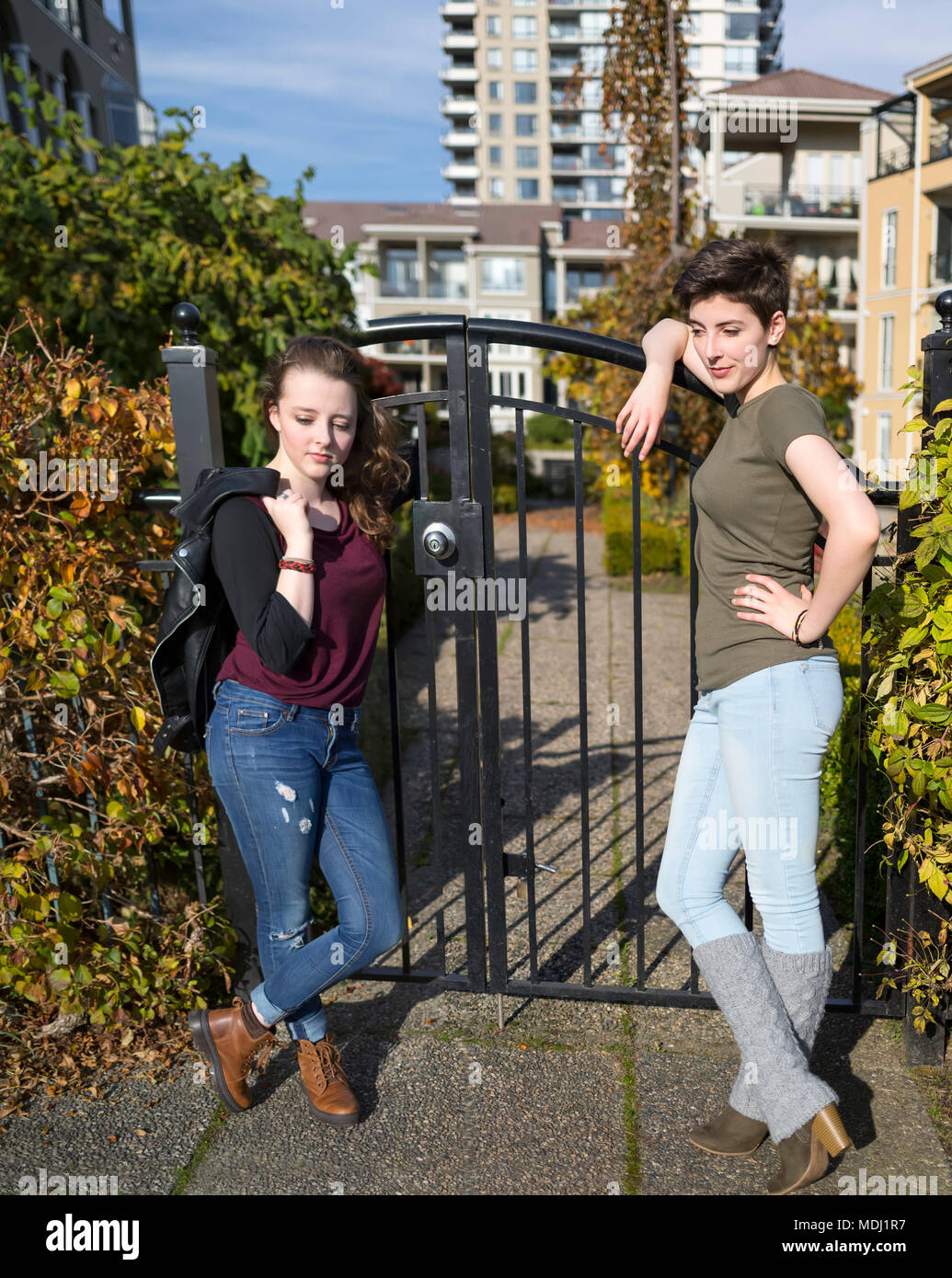 Two young women stand leaning against a metal gate looking confident and cool with housing in the background Stock Photo