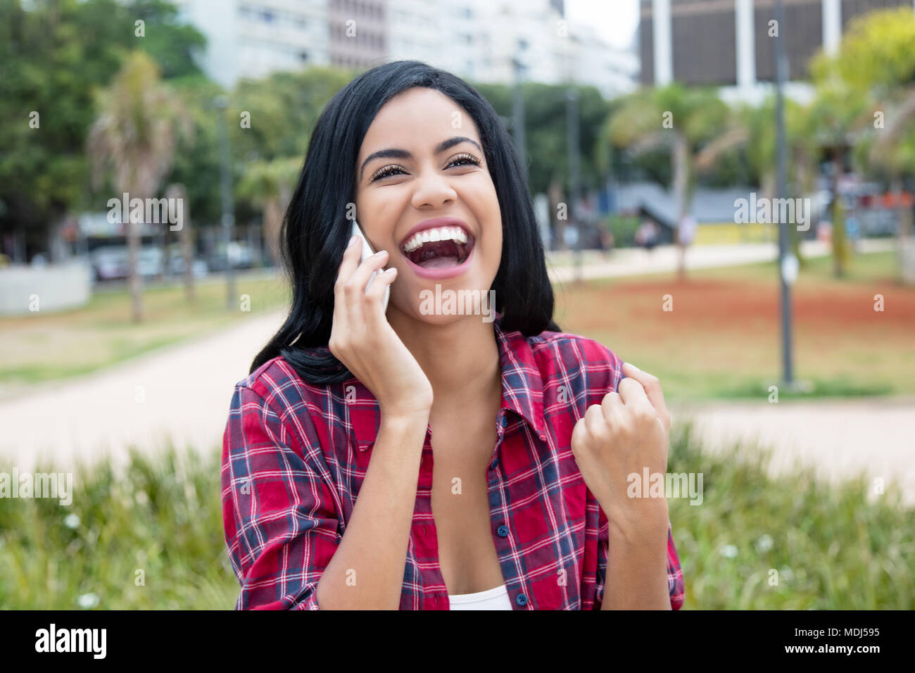 Cheering latin american young adult woman at mobile phone outdoors in the city Stock Photo