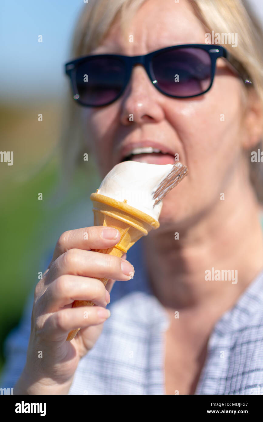 Woman wearing sunglasses holding and licking a vanilla ice cream in a cone.with a chocolate flake in it. Soft focus on her face. Stock Photo