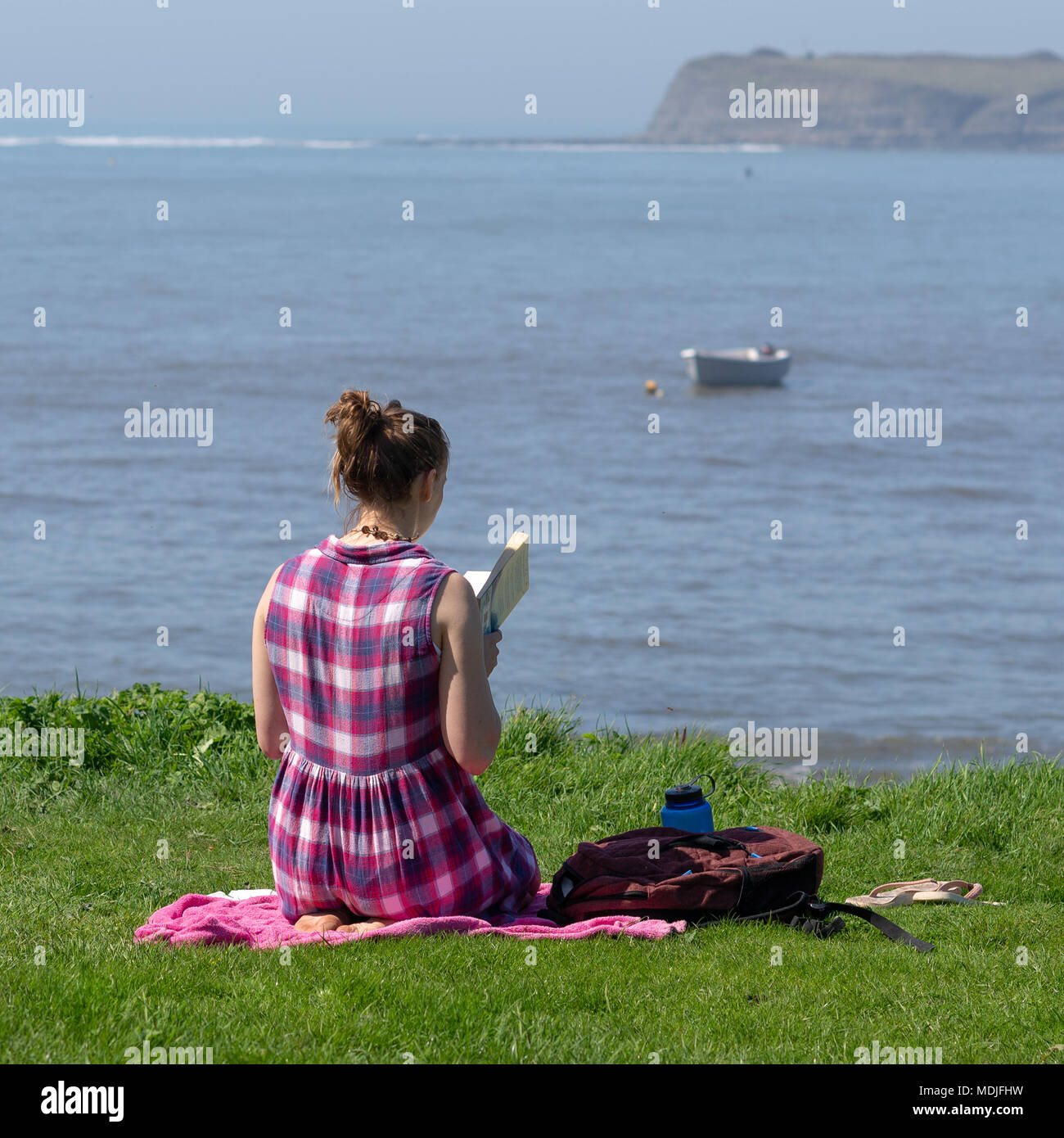 A young woman in a checked dress sitting on a blanket on a grassy cliff overlooking the sea and reading a book in sunny weather. Stock Photo