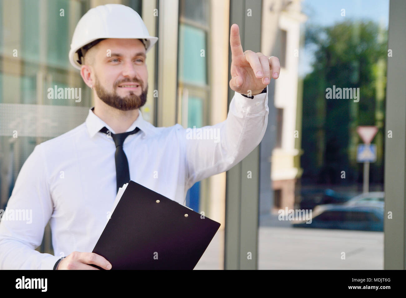 A man working in a white helmet and white shirt with a tablet in his hands clicks his index finger in the virtual screen. Engineer. Planning of constr Stock Photo
