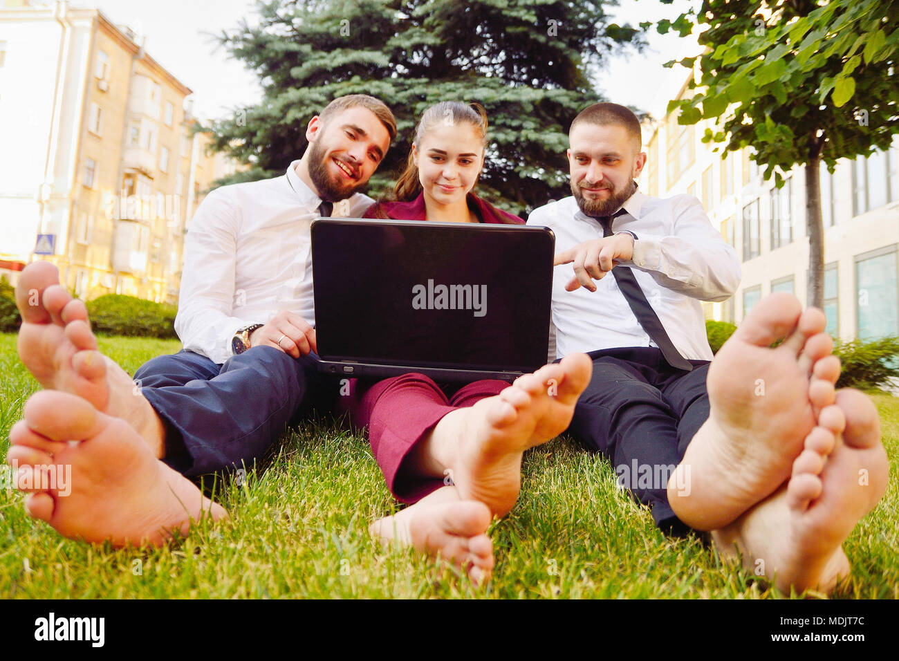 Young office workers with bare feet sit on a green lawn with a laptop