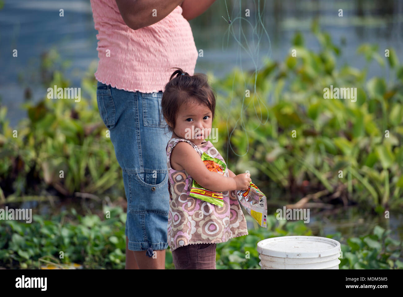 A young, indigenous Itza Maya girl eats candy while her mother fishes in Lake Peten, northern Guatemala. Stock Photo