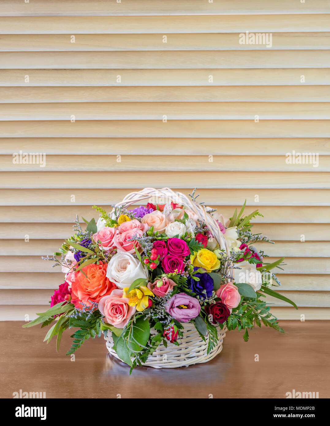 beautiful bouquet of flowers, colorful roses with green leaves standing in a wicker basket on a brown table, light wooden blinds in the background, re Stock Photo