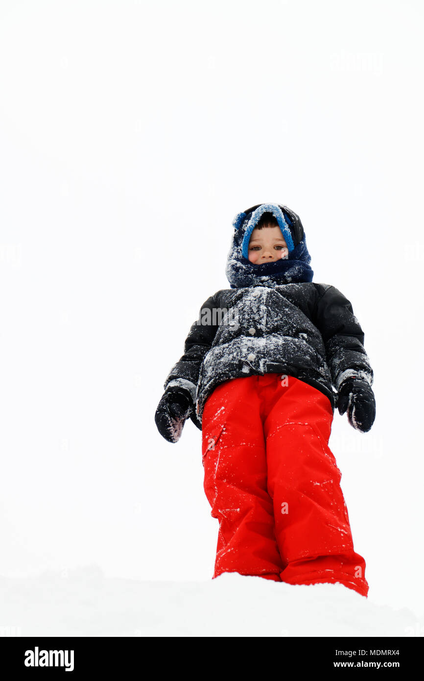 Looking up at a young boy (5 yr old) dressed in outdoor winter clothes. He is stood on top of a pile of snow with an almost white sky behind. Stock Photo
