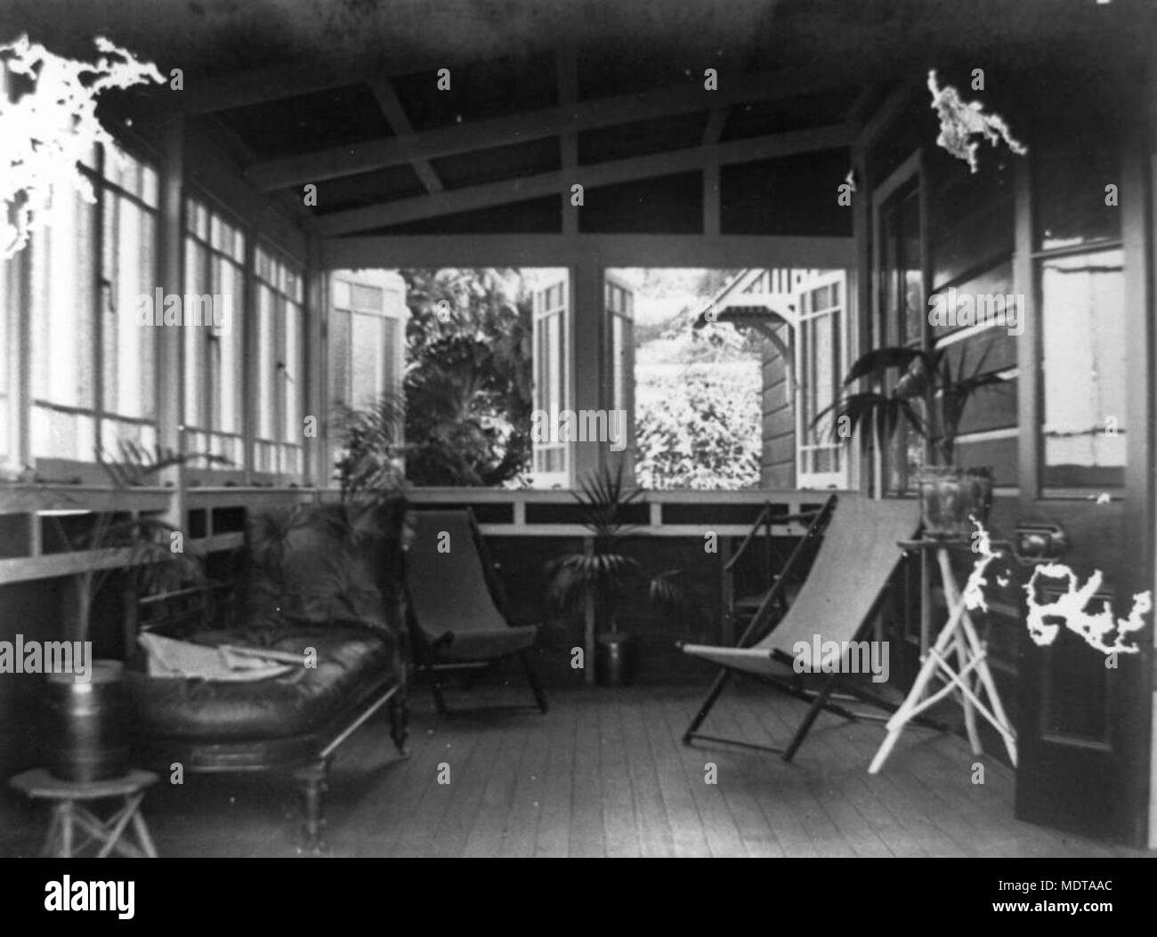 Furnishings on a verandah of a Queensland home, ca 1925. Description: A covered verandah with open windows, furnished with deck chairs, a chaise lounge and pot plants. Stock Photo