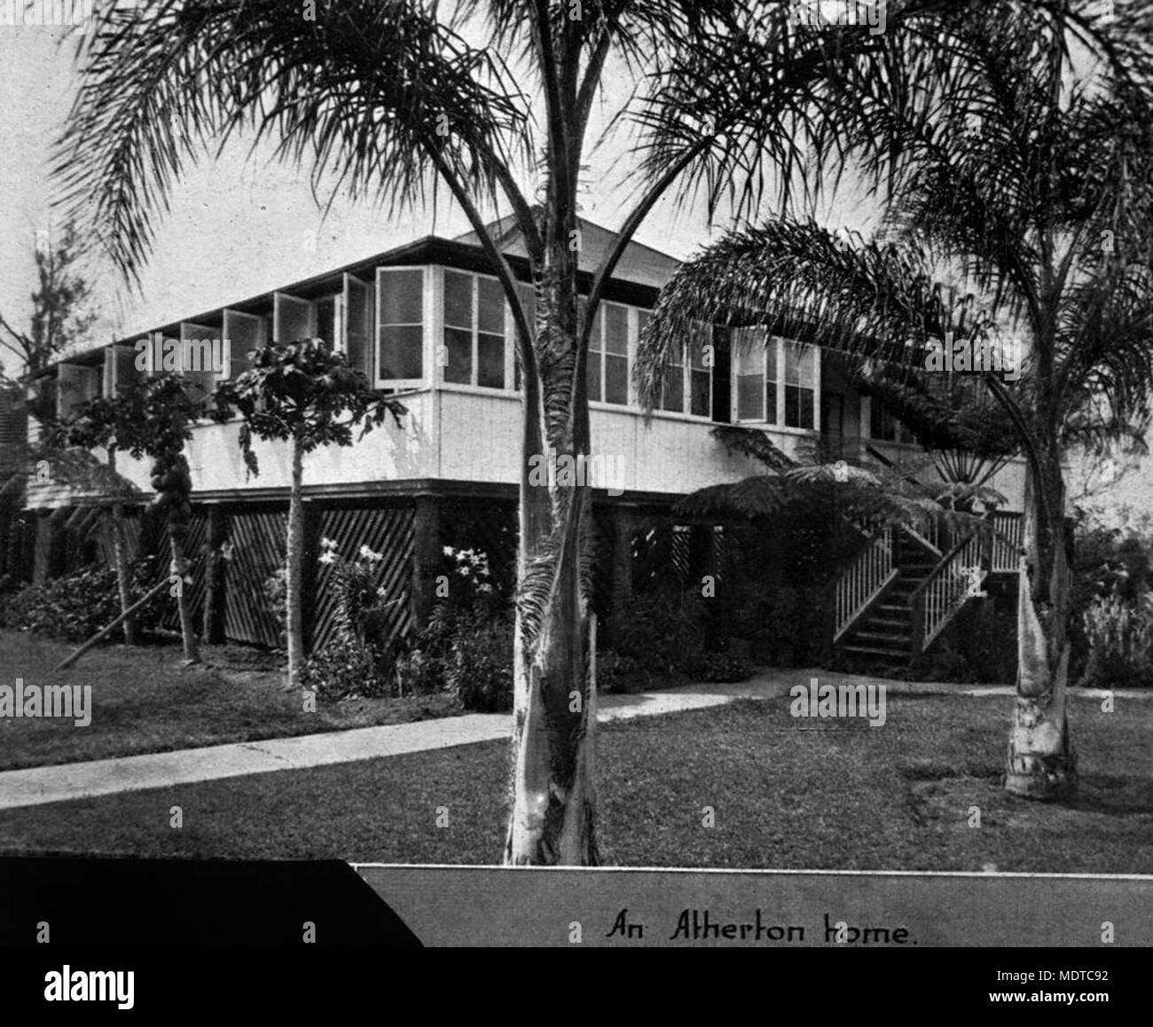 Queenslander home set in a tropical garden at Atherton, Queensland,. Location: Atherton, Queensland    Description: This highset house in Atherton has enclosed verandahs, approached from the front by divided stairs. The windows on the verandah are open to catch the cool breezes.  Atherton was named after John Atherton, the pioneer pastoralist, who brought cattle into the district from Rockhampton in 1877. He took up Emerald End Station on the banks of the Barron River. Settlement in Atherton began with the development of a cedar logging camp known as Prior's Pocket. Atherton is the agricultura Stock Photo