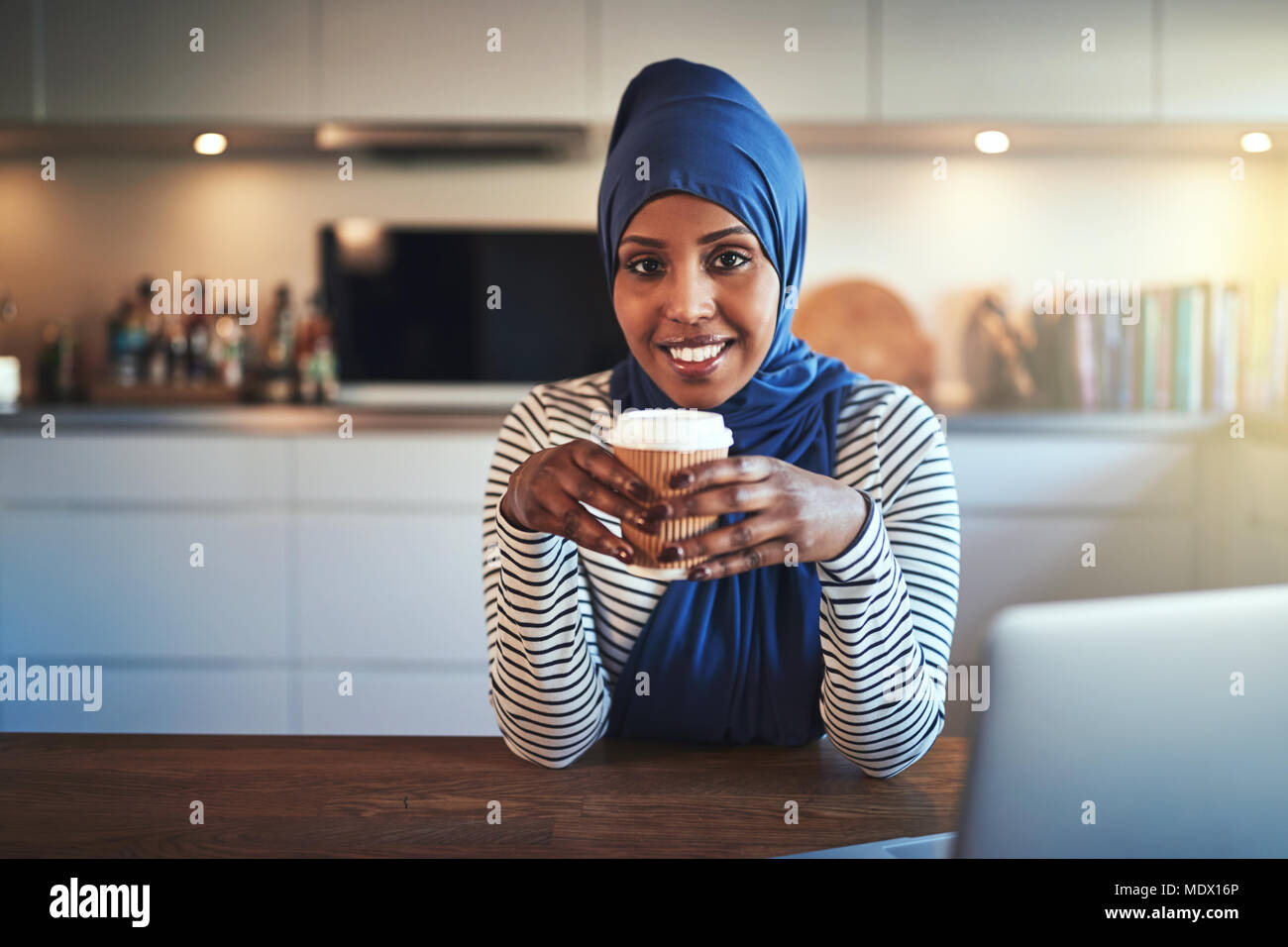 Smiling young Arabic woman wearing a hijab sitting in her kitchen at ...