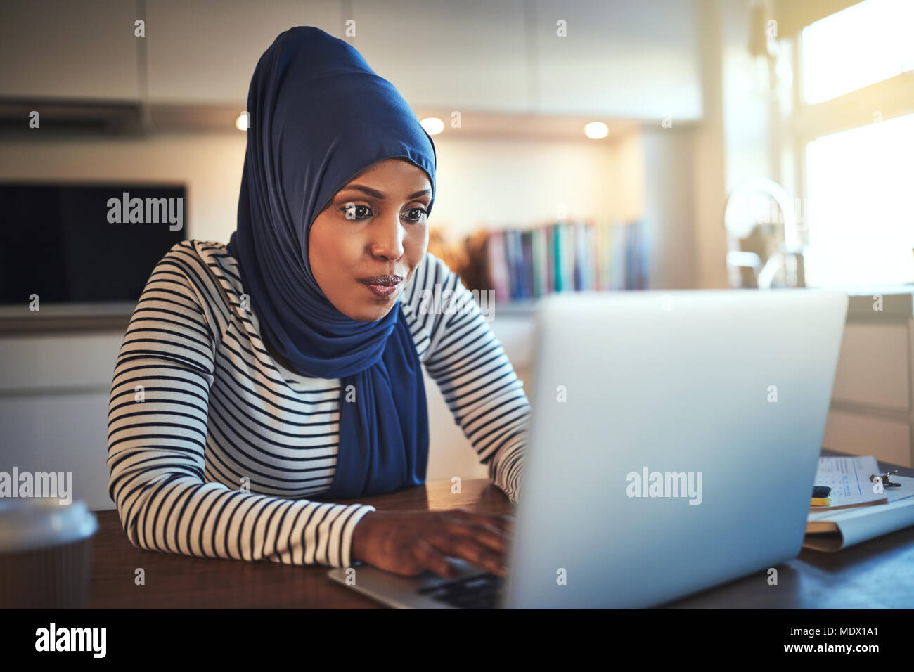 Young Arabic woman wearing a hijab sitting at her kitchen table using a ...