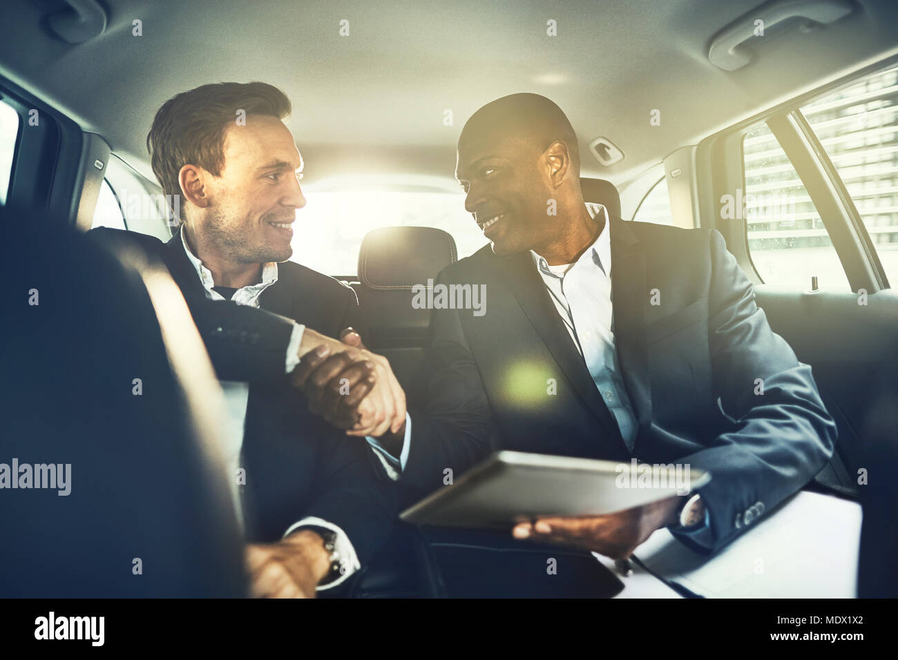 Two smiling businessman shaking hands together while sitting in the backseat of a car driving through the city Stock Photo