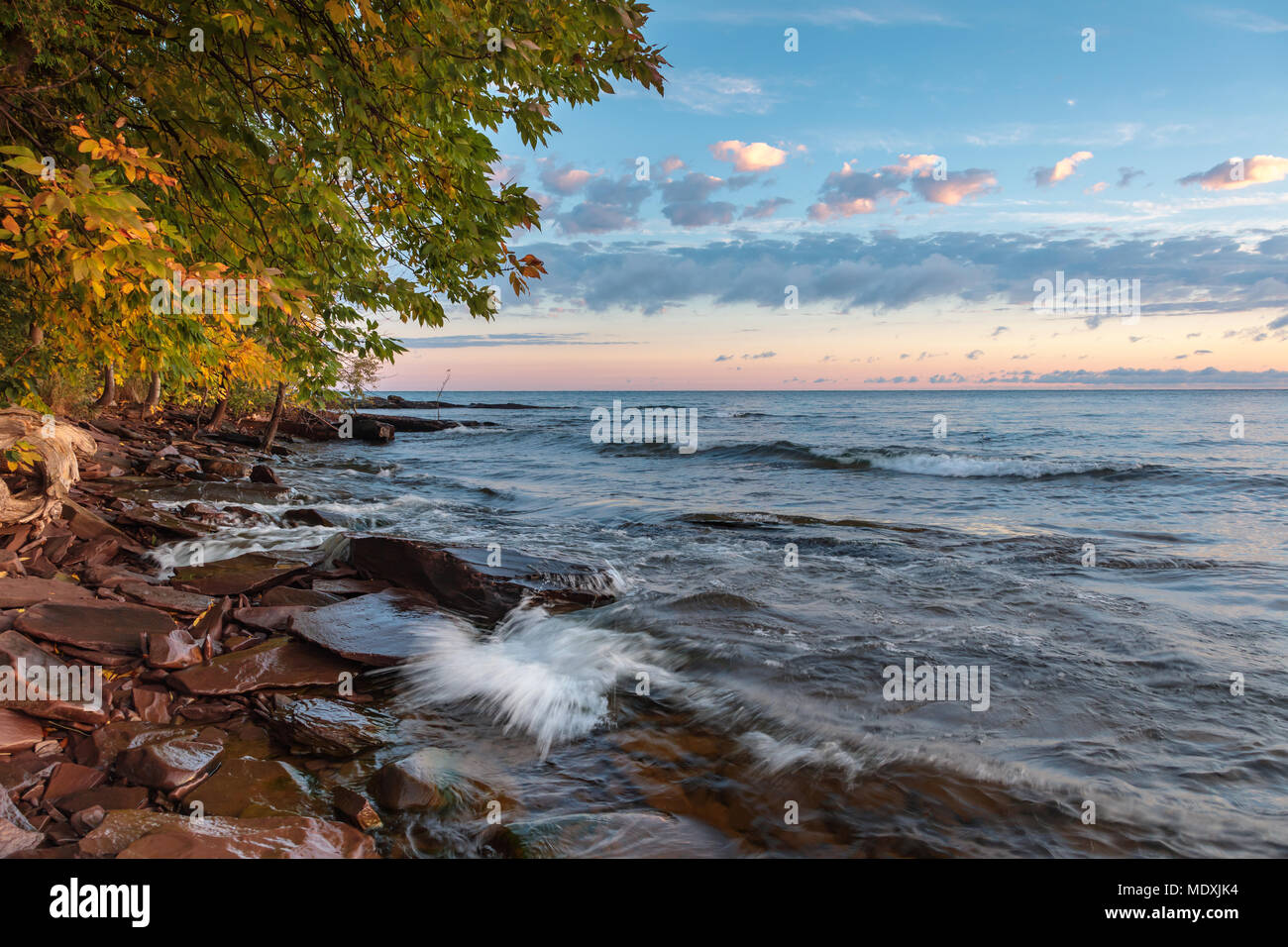 Early morning sun lights up sandstone rocks and tree branches as Lake Superior waves roll up to the shoreline. Puffy clouds are in the sky above Stock Photo