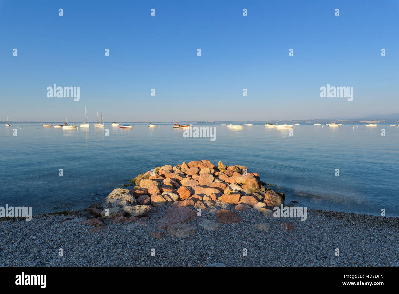 Rocks piled on beach at shoreline of Lake Garda (Lago di Garda) with boats anchored in the distance, Bardolino in Veneto, Italy Stock Photo