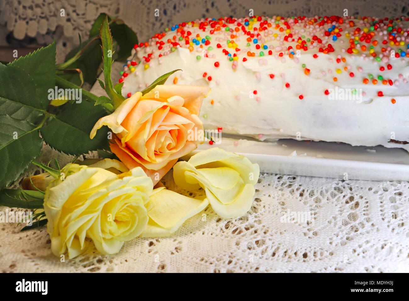 A white cake decorated with colored candies on a tray on a table with a lace tablecloth and roses Stock Photo