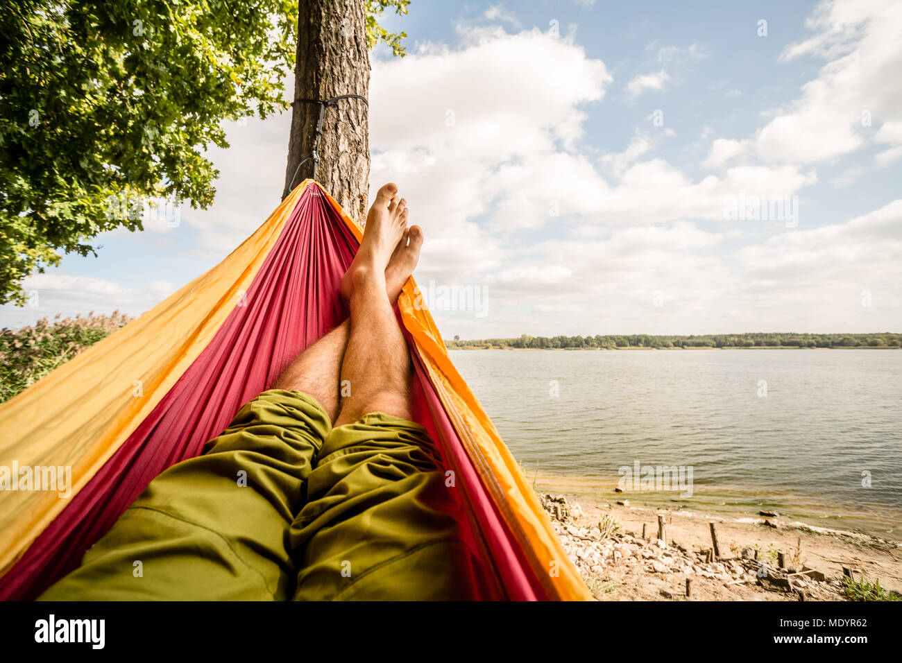 Relaxing in the hammock at the beach under a tree, summer day. Barefoot man laying in hammock, looking on a lake, inspiring landscape Stock Photo