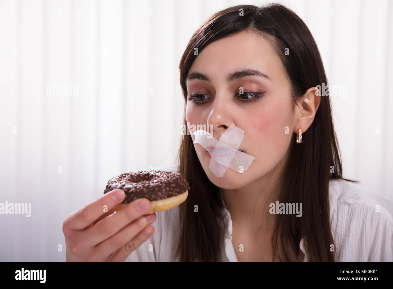 Close-up Of A Young Woman With Sticky Tape Over Her Mouth Unable To Eat Donut Stock Photo