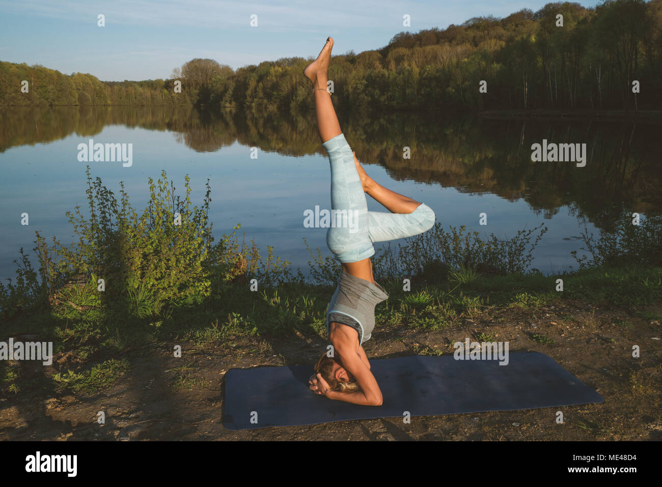 Young woman exercising yoga by the lake at sunset, people travel nature wellbeing and relaxation concept. Shot in France, Europe. HEADSTAND Stock Photo