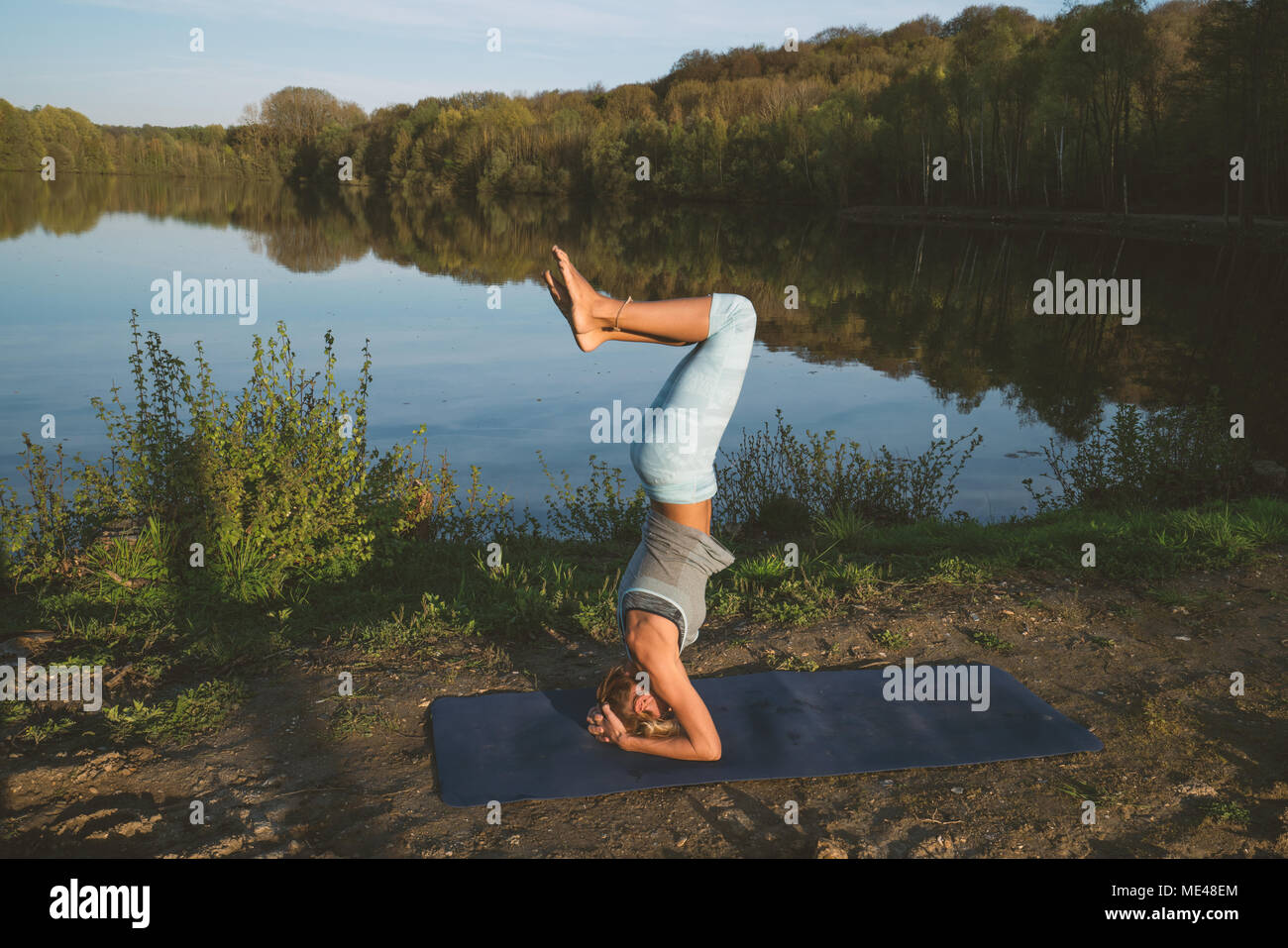 Young woman exercising yoga by the lake at sunset, people travel nature wellbeing and relaxation concept. Shot in France, Europe. HEADSTAND Stock Photo