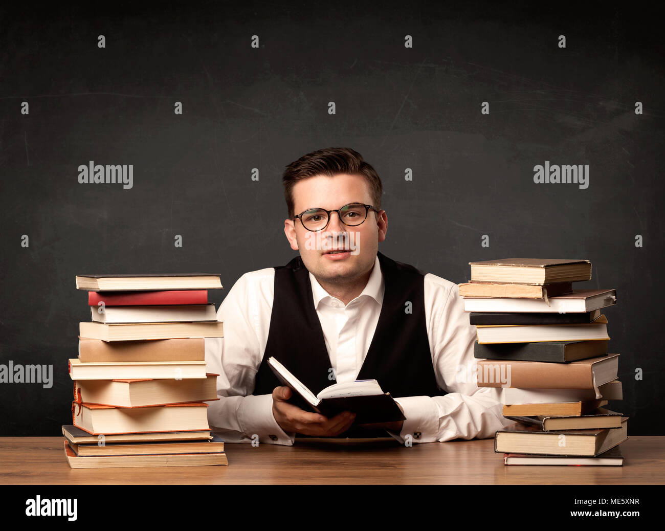 A young teacher in glasses sitting at classroom desk with pile of books ...