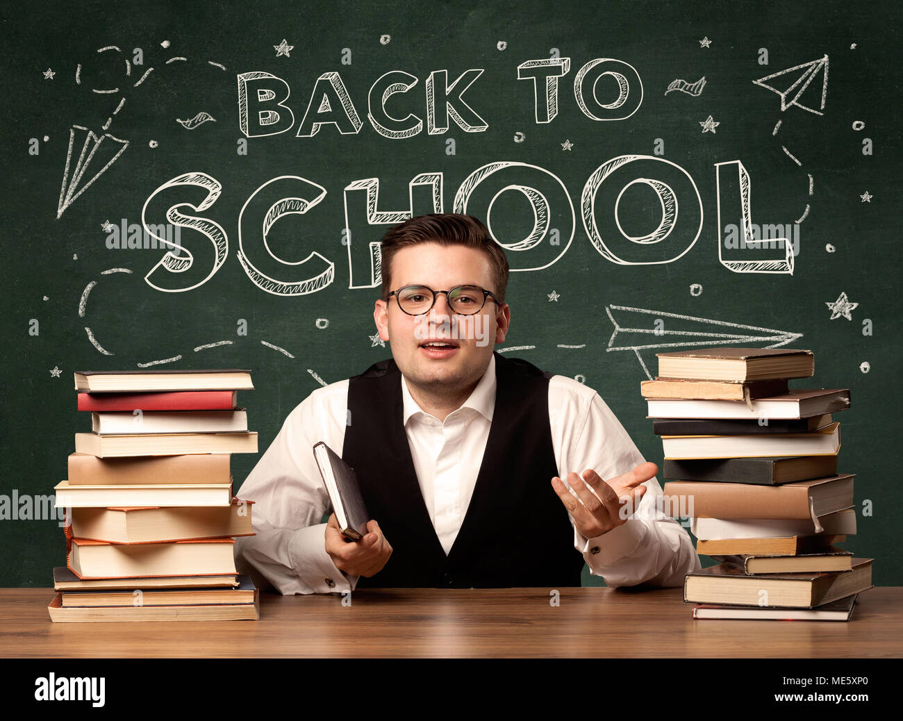 A young teacher in glasses sitting at classroom desk with pile of books ...