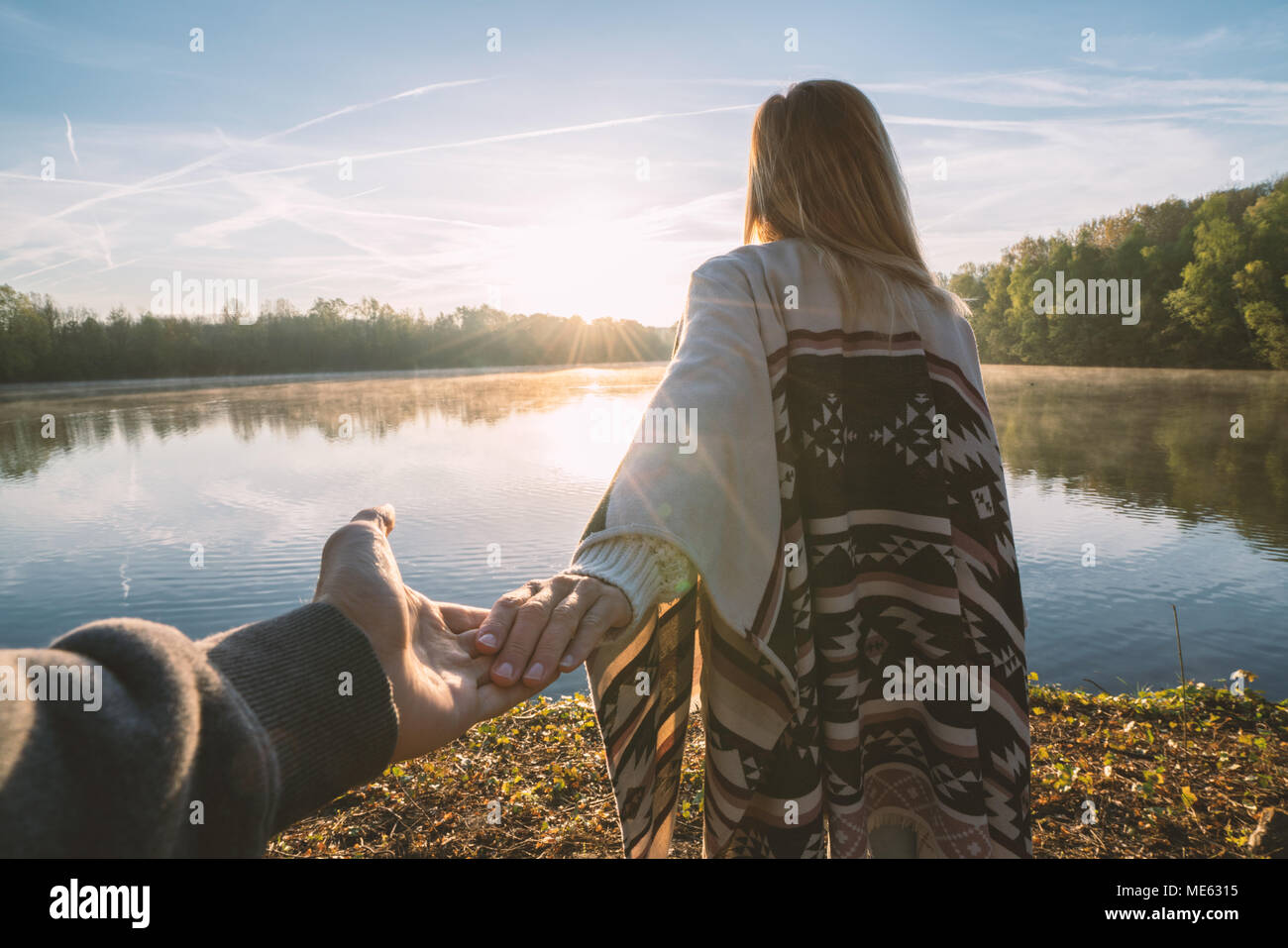Follow me to concept, Young woman leading boyfriend to the lakeshore at sunrise, holding hands, People travel couple beginning. Shot in France, Europe Stock Photo