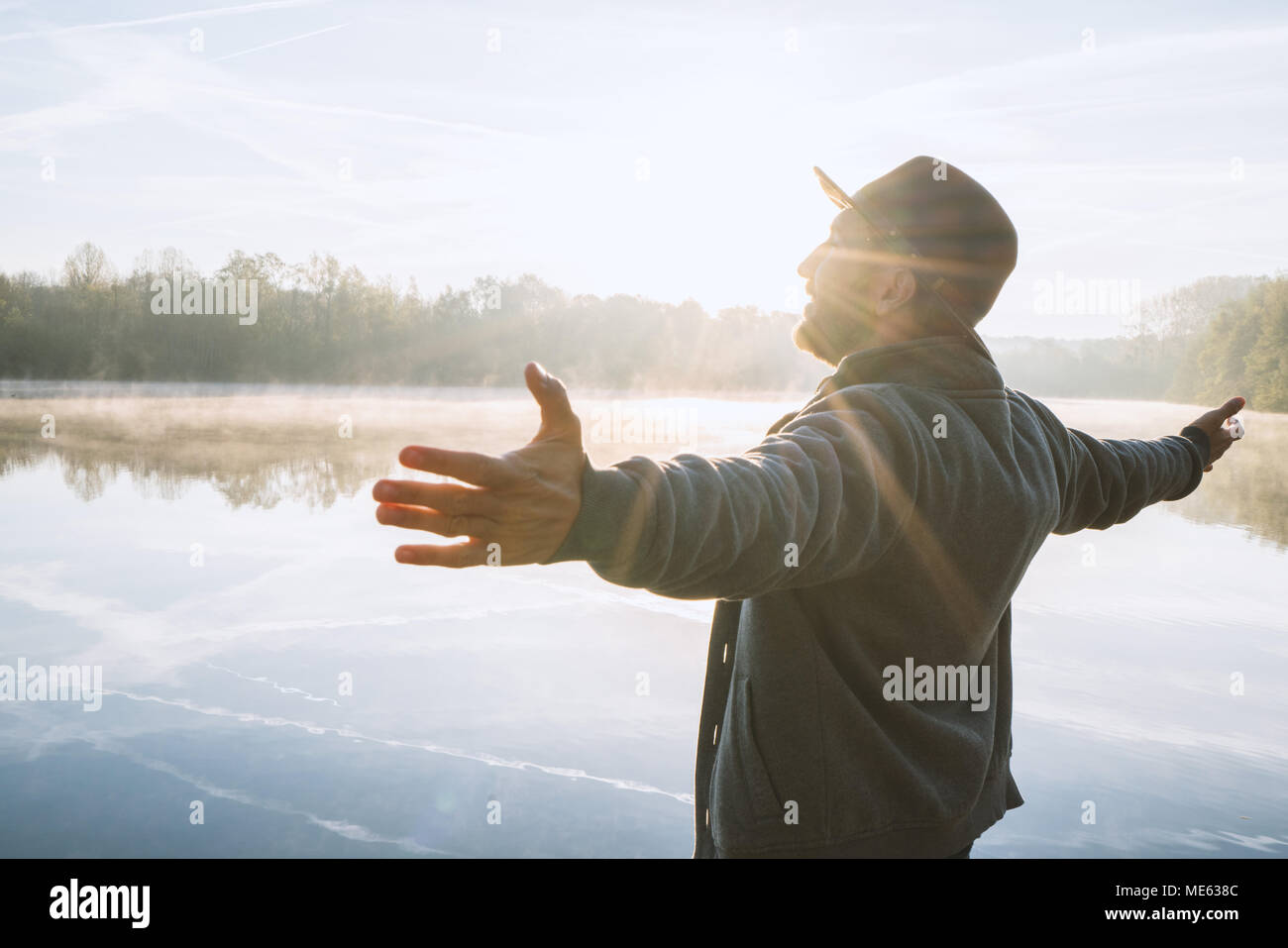 Young man arms outstretched by the lake at sunrise enjoying freedom and life, people travel wellbeing concept. Shot in France, Europe. Stock Photo