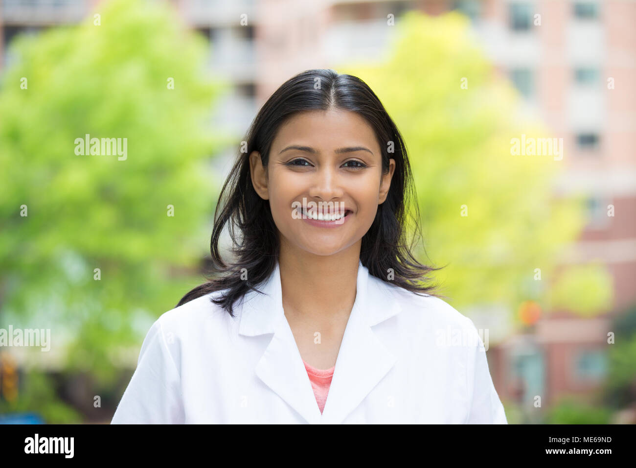 Closeup portrait of confident, smiling female health care professional in white lab coat, isolated background of blurred trees and buildings Stock Photo