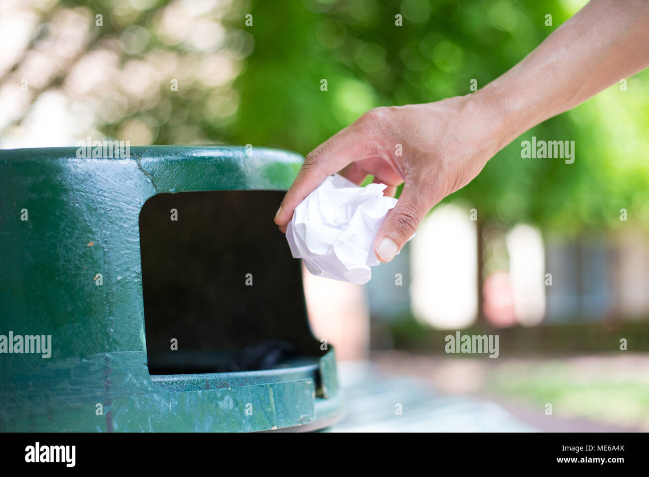 Closeup cropped portrait of someone tossing crumpled piece of paper in trash can, isolated outdoors green trees background Stock Photo