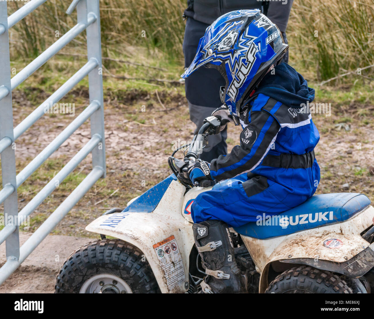 West Linton, Scottish Borders, Scotland, United Kingdom, April 21st 2018.  Spring sunshine in the countryside, with a young boy wearing a motorcycle helmet driving a small Suzuki quad bike on a farm track Stock Photo