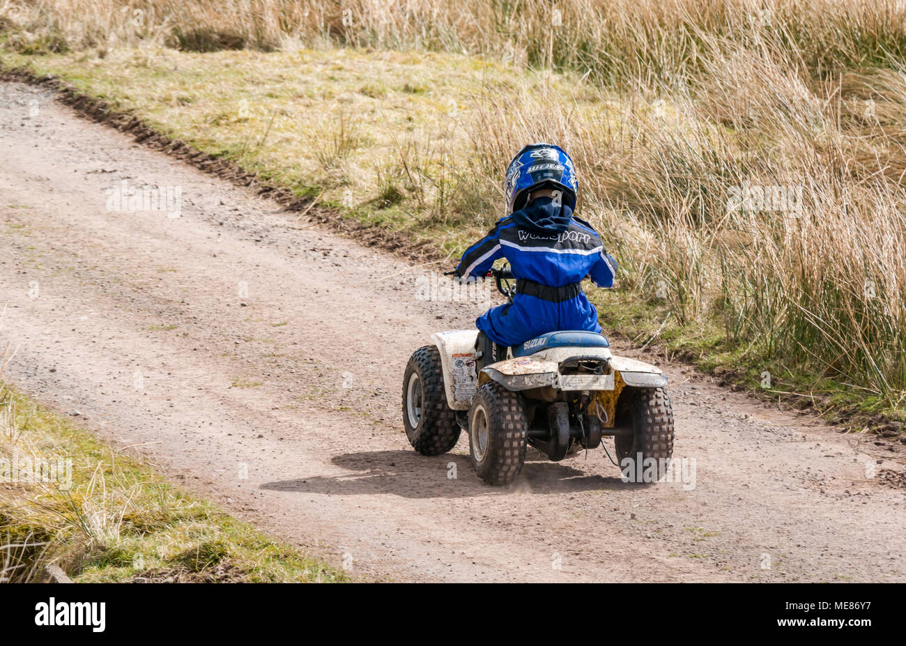 West Linton, Scottish Borders, Scotland, United Kingdom, April 21st 2018.  Spring sunshine in the countryside, with a young boy wearing a motorcycle helmet driving a small quad bike on a farm track Stock Photo