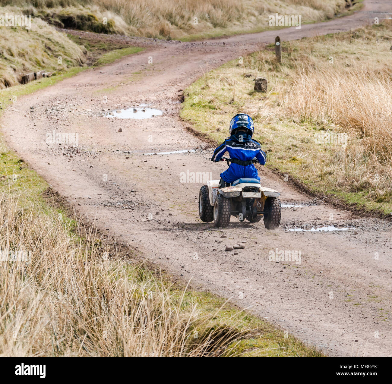 West Linton, Scottish Borders, Scotland, United Kingdom, April 21st 2018.  Spring sunshine in the countryside, with a young boy wearing a motorcycle helmet driving a small quad bike on a farm track Stock Photo