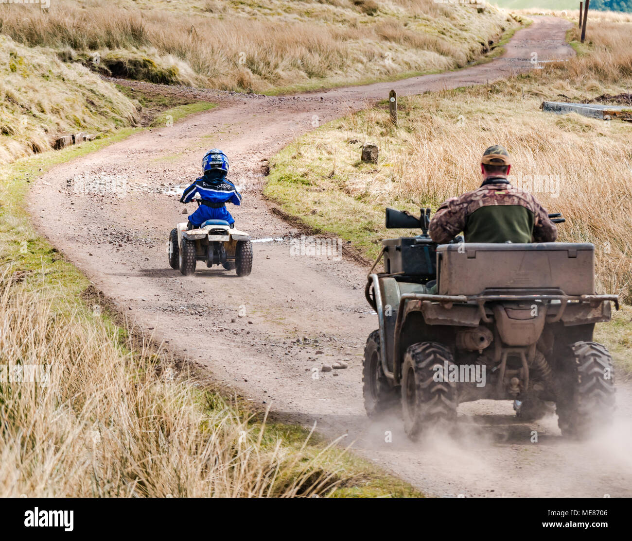 West Linton, Scottish Borders, Scotland, United Kingdom, April 21st 2018.  UK Weather: Warm Spring sunshine in the countryside, with a young boy wearing a motorcycle helmet driving a small quad bike on a farm track, followed by his farmer father on an all terrain vehicle with a shotgun resting on the bonnet Stock Photo