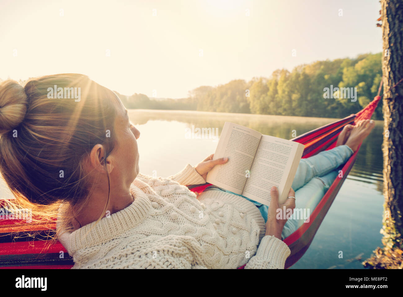 Young woman relaxing on hammock by the lake at sunrise reading a book ...