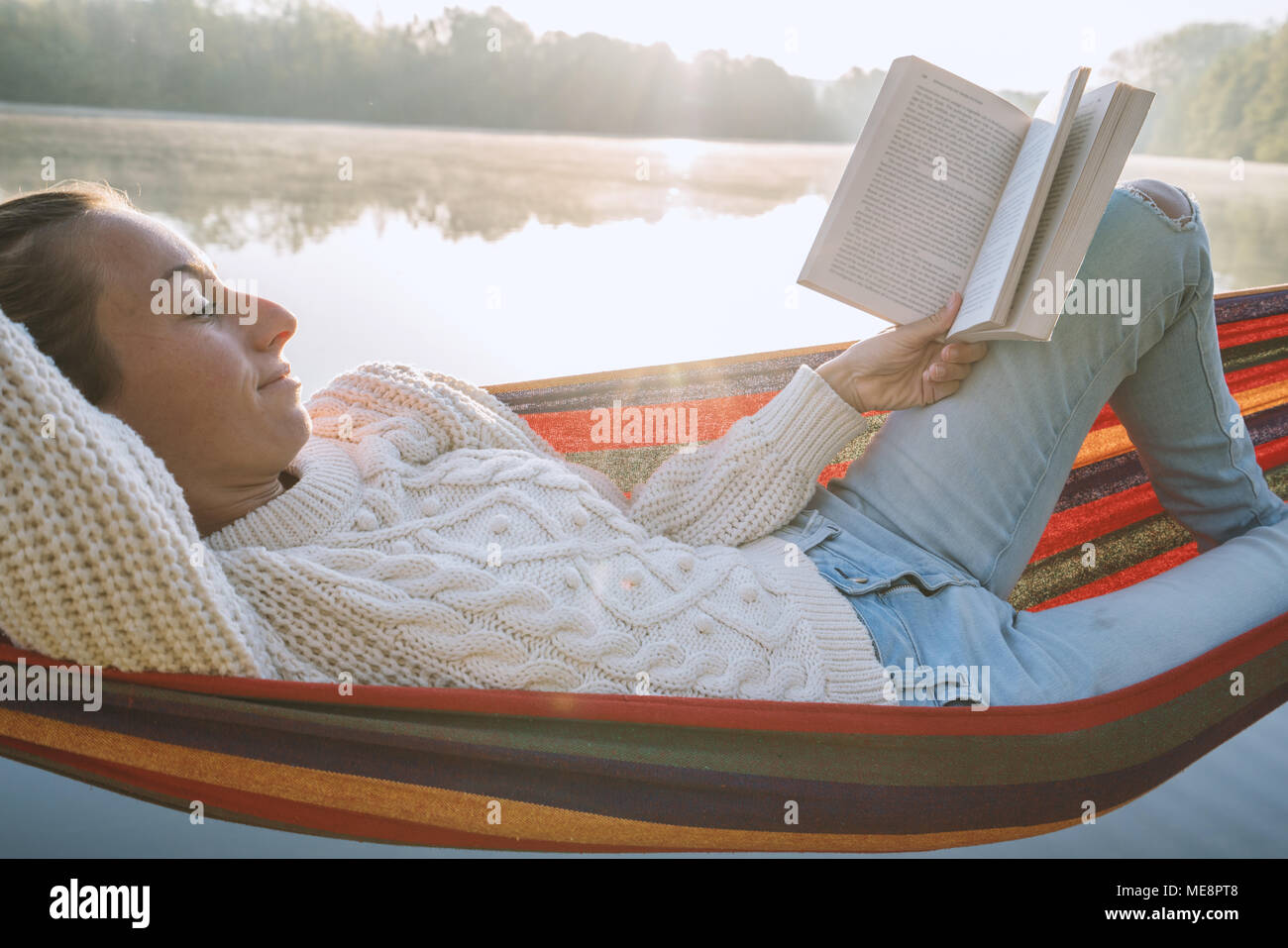 Young woman relaxing on hammock by the lake at sunrise reading a book. People travel relaxation wellbeing learning concept. France, Europe Stock Photo