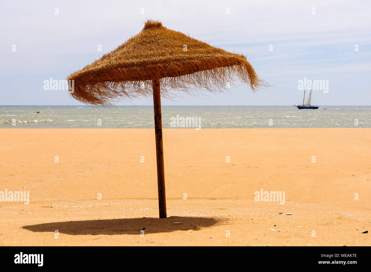 Straw parasol on a sandy beach with a yacht sailing past Stock Photo