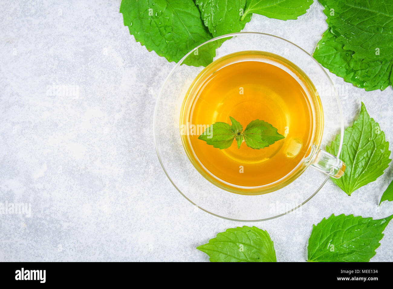 Leaves of fresh green nettle and a clear glass cup of herbal nettle tea on a gray concrete table. Top view Stock Photo