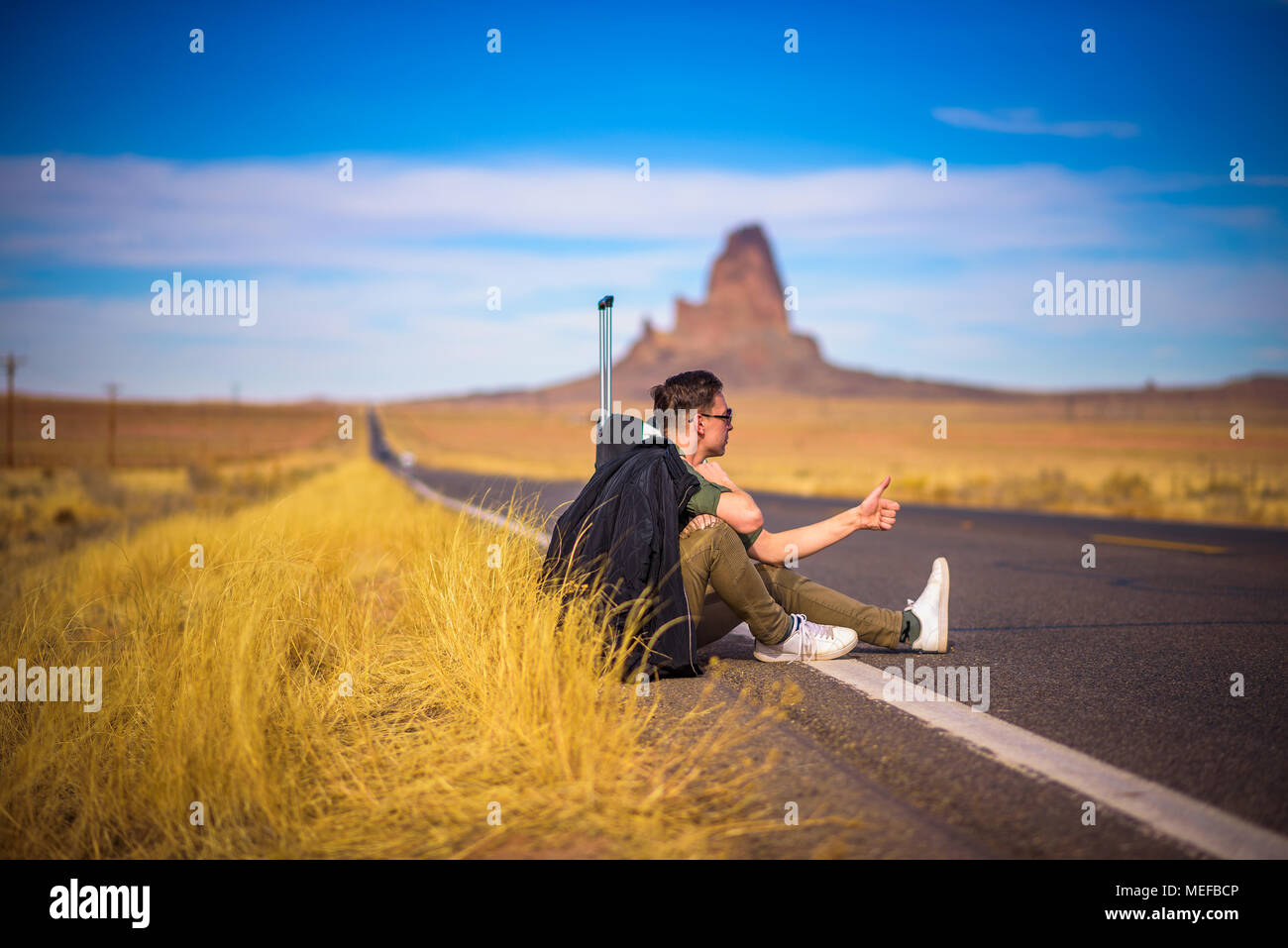 Tired hitch-hiker with suitcase sitting on a road Stock Photo