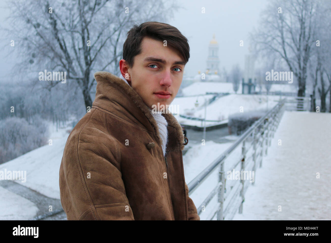 Young handsome european guy in warm coat and scarf on the snow-covered bridge during cold winter Stock Photo