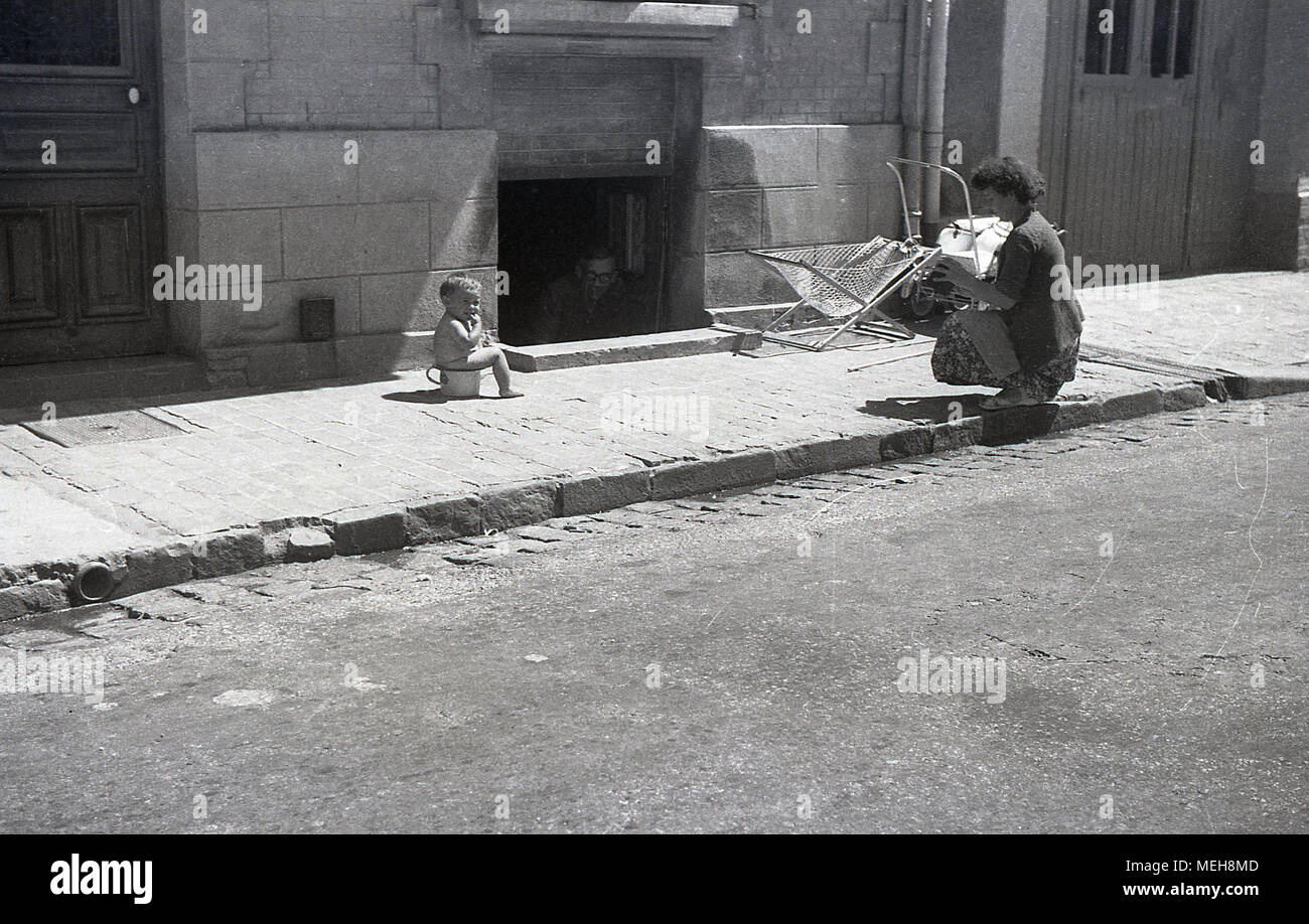 1930s, summertime in the port of Caen, Normandy, France, mother kneeling down about to take a picture of her baby sitting on a potty in the middle of the pavement, while a man in a semi-basement room looks on. Stock Photo