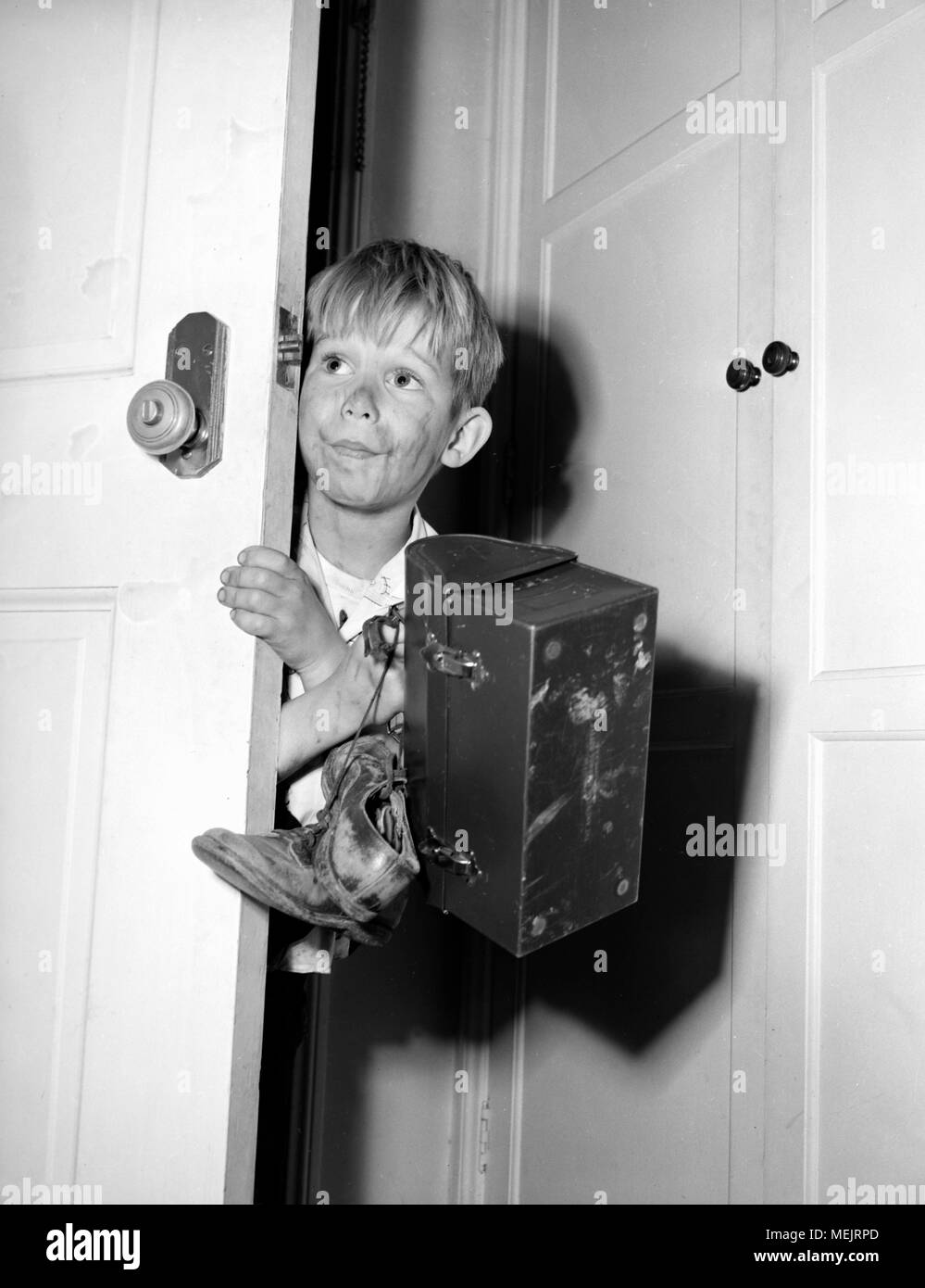 A young boy warily comes in the house with his dirty clothes, muddy shoes, and his lunch box after school, ca. 1964. Stock Photo