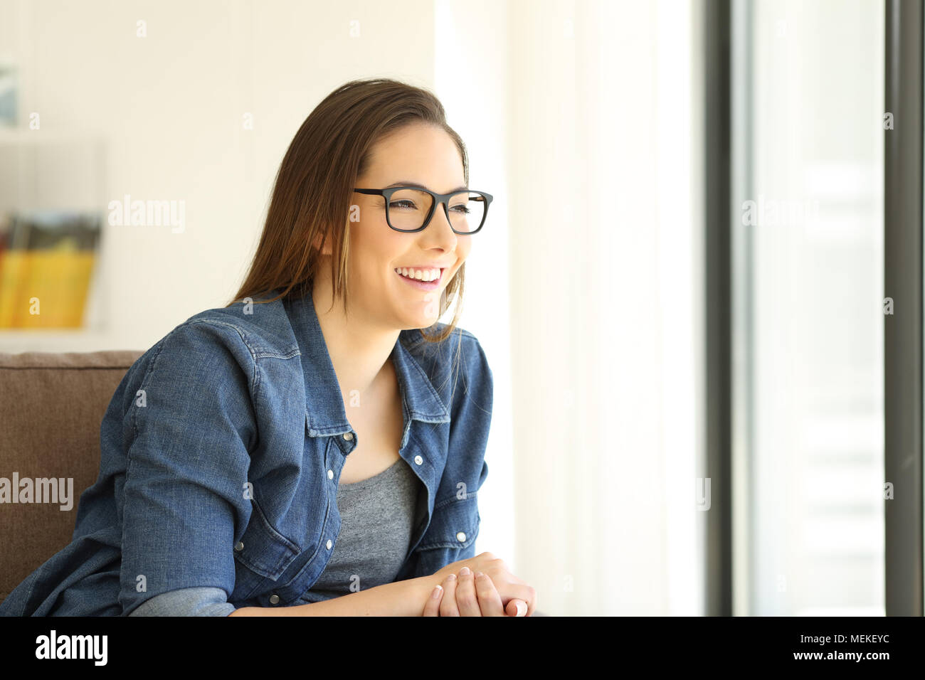 Happy girl wearing eyeglasses looking through a window sitting on a couch in the living room at home Stock Photo