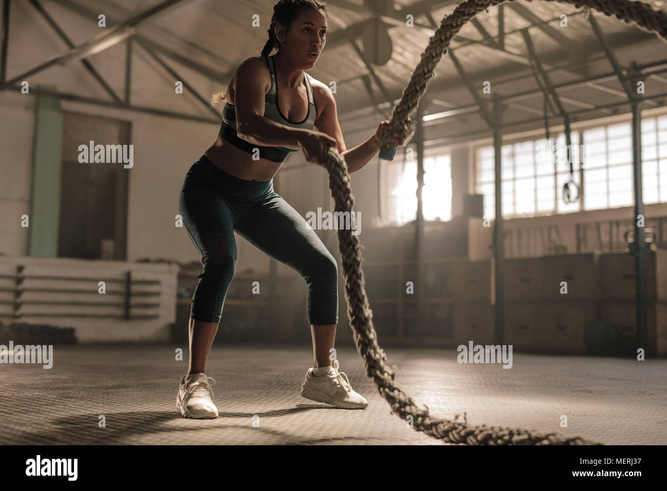 Young woman doing strength training using battle ropes at the gym. Athlete moving the ropes in wave motion as part of fat burning workout. Stock Photo