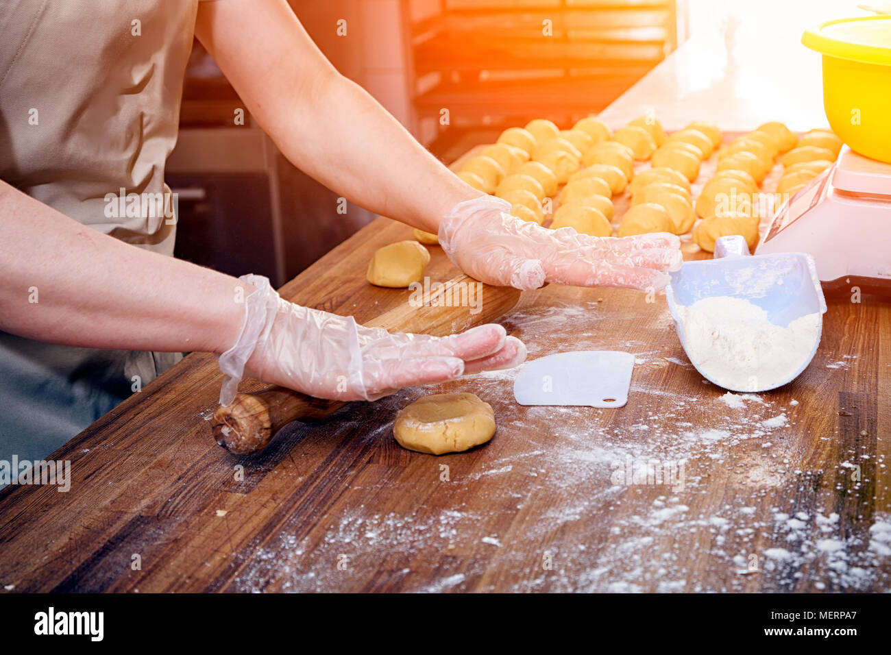 A close-up of a female baker in protective gloves pulls out the dough for cooking buns on a knotted table with a rolling pin Stock Photo