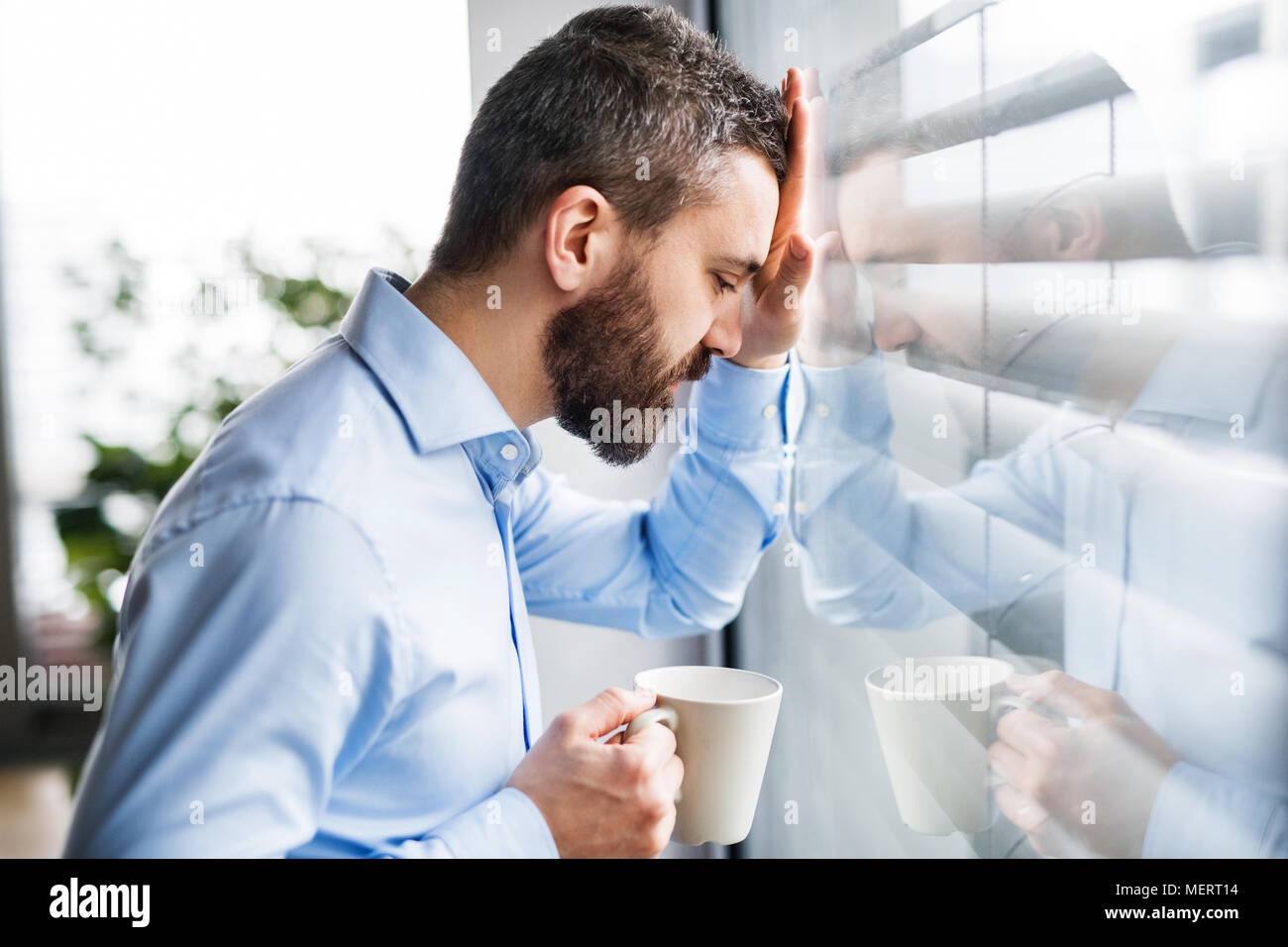 An unhappy man by the window holding a cup of coffee. Stock Photo