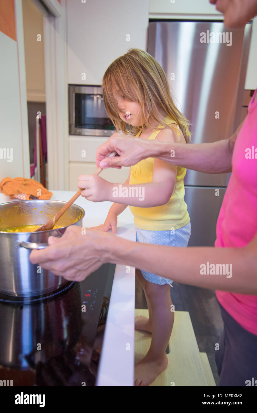 Four years old blonde child on stool o ladder helping her woman mother to cook, together as a team, in electrical cooktop with a saucepan, in the kitc Stock Photo