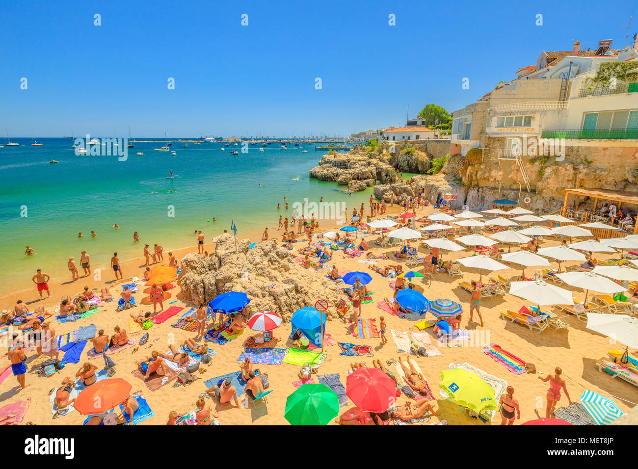 Cascais, Portugal - August 6, 2017: people sunbathing on Praia da Rainha, a small beach with cliffs in Cascais center, Lisbon Coast. Turquoise sea in summer holidays. Fishing Harbour on background. Stock Photo